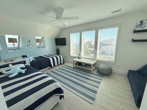 A bedroom with two beds, a ceiling fan, a TV, a window with blinds, and a view of the city. - Blue Horizon - Oceanfront House on Harbor Island (Harbor Island)