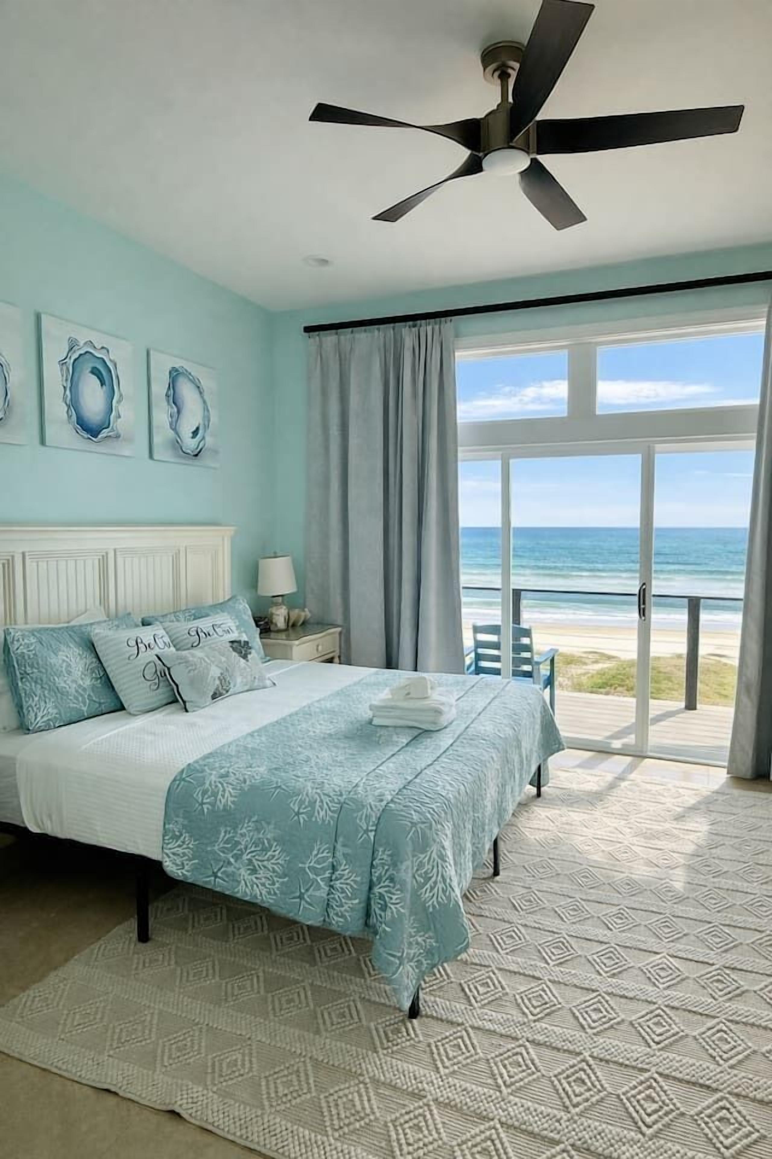 A bedroom with a bed, a ceiling fan, a nightstand, a lamp, and a view of the ocean through a sliding door.