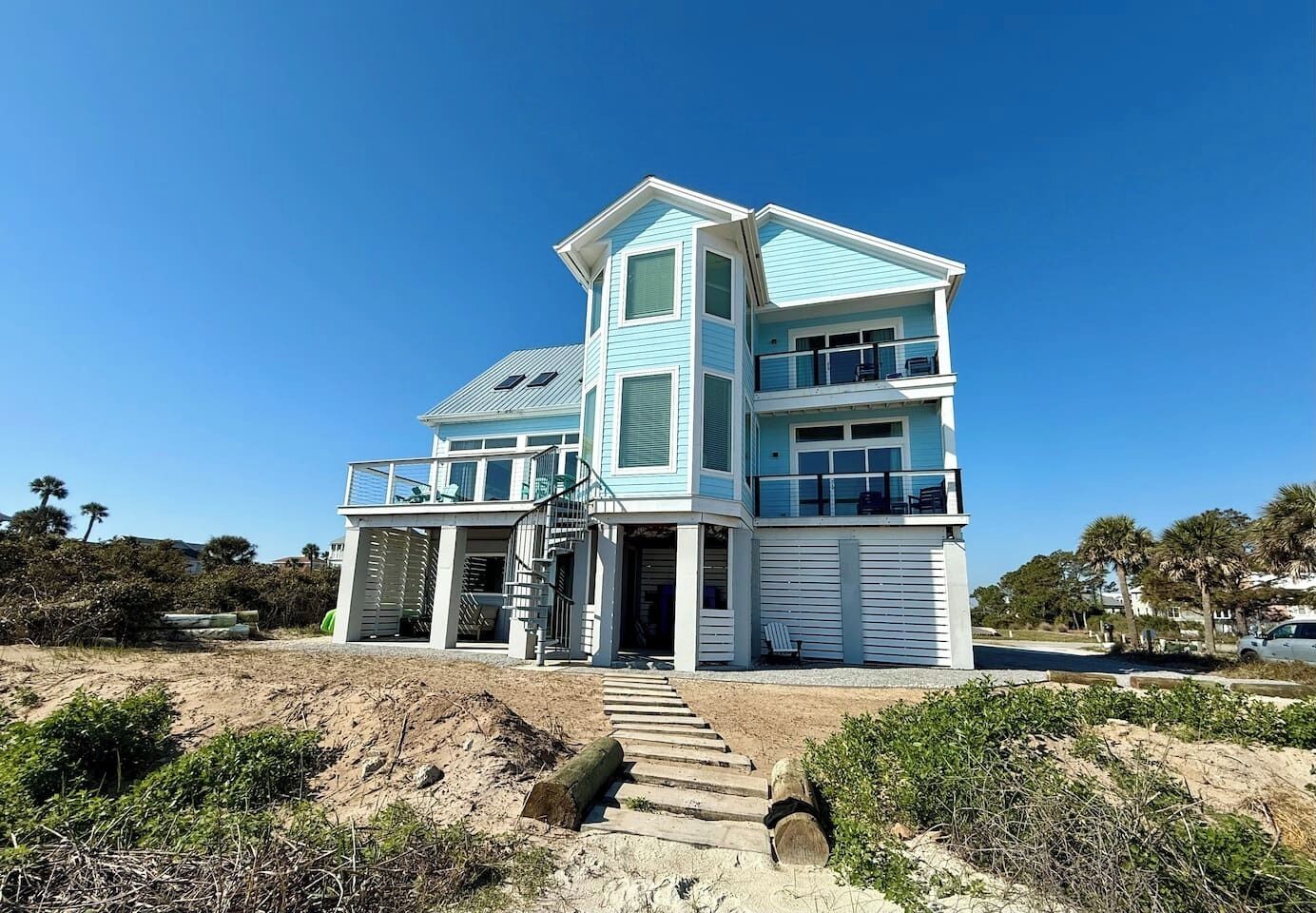 A multi-story beachfront house with a prominent balcony, a staircase leading to the entrance, and a sandy foreground.