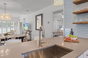 A modern kitchen with a central island, stainless steel sink, and a dining area with a white table and chairs. - New Beach House Rental. Welcome to your Isle of Palms Oasis! (Isle Of Palms)