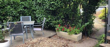 A patio area with a metal table and chairs, a hedge, and a planter with red flowers.