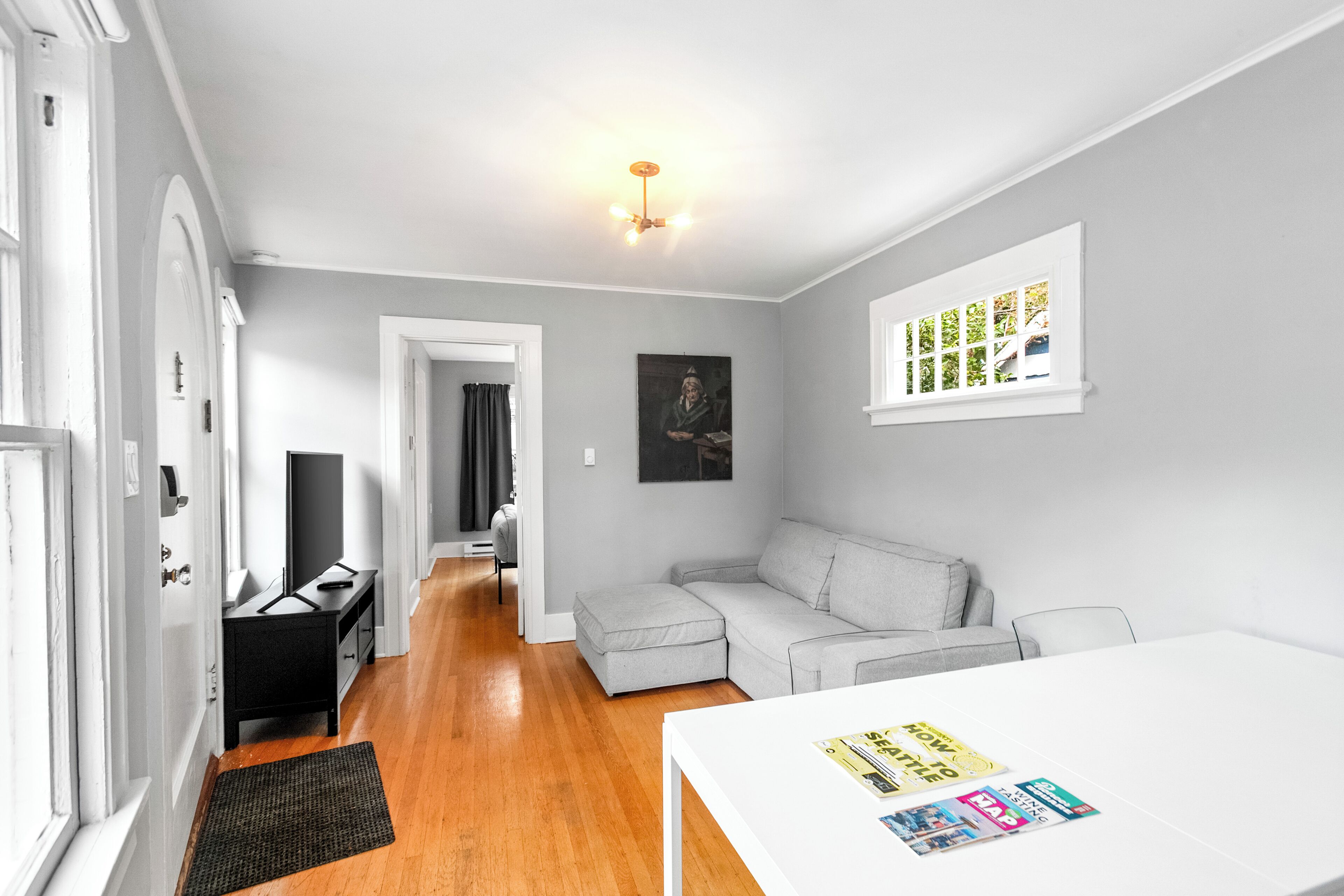 A modern living room with a grey sofa, a white table with magazines, a black TV stand, and a door leading to another room.