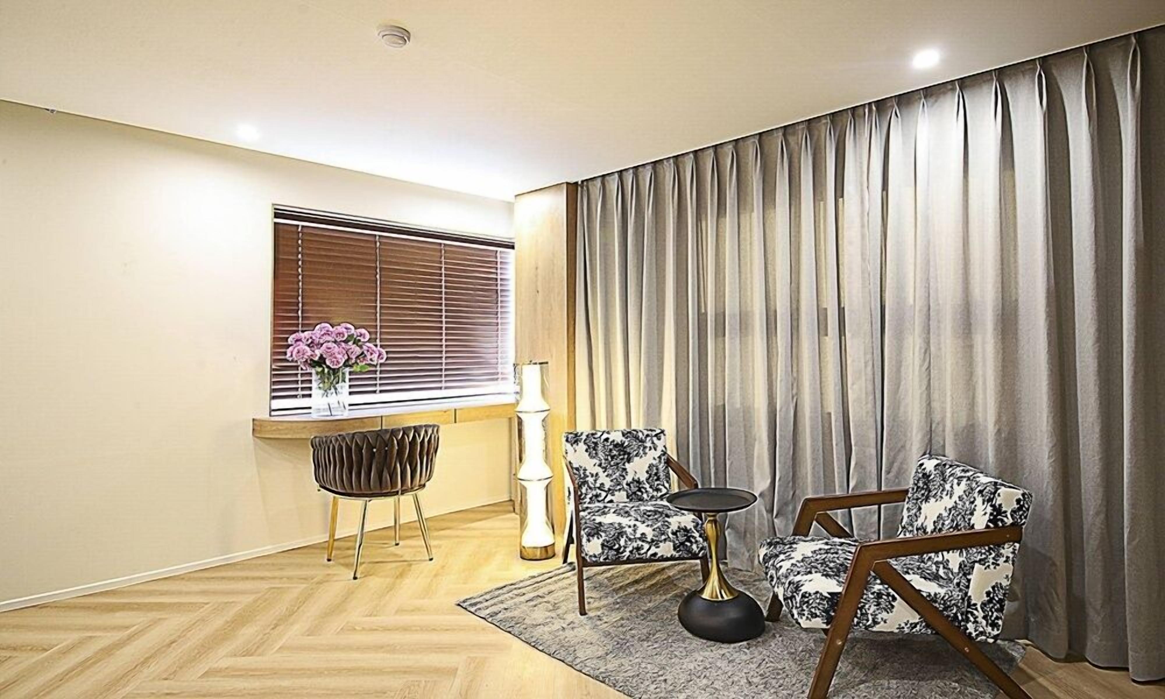 Una habitación con piso de madera, una ventana con persianas, un jarrón con flores, una lámpara, dos sillones estampados y un gran cortinaje.