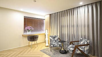 Una habitación con piso de madera, una ventana con persianas, un jarrón con flores, una lámpara, dos sillones estampados y un gran cortinaje.
