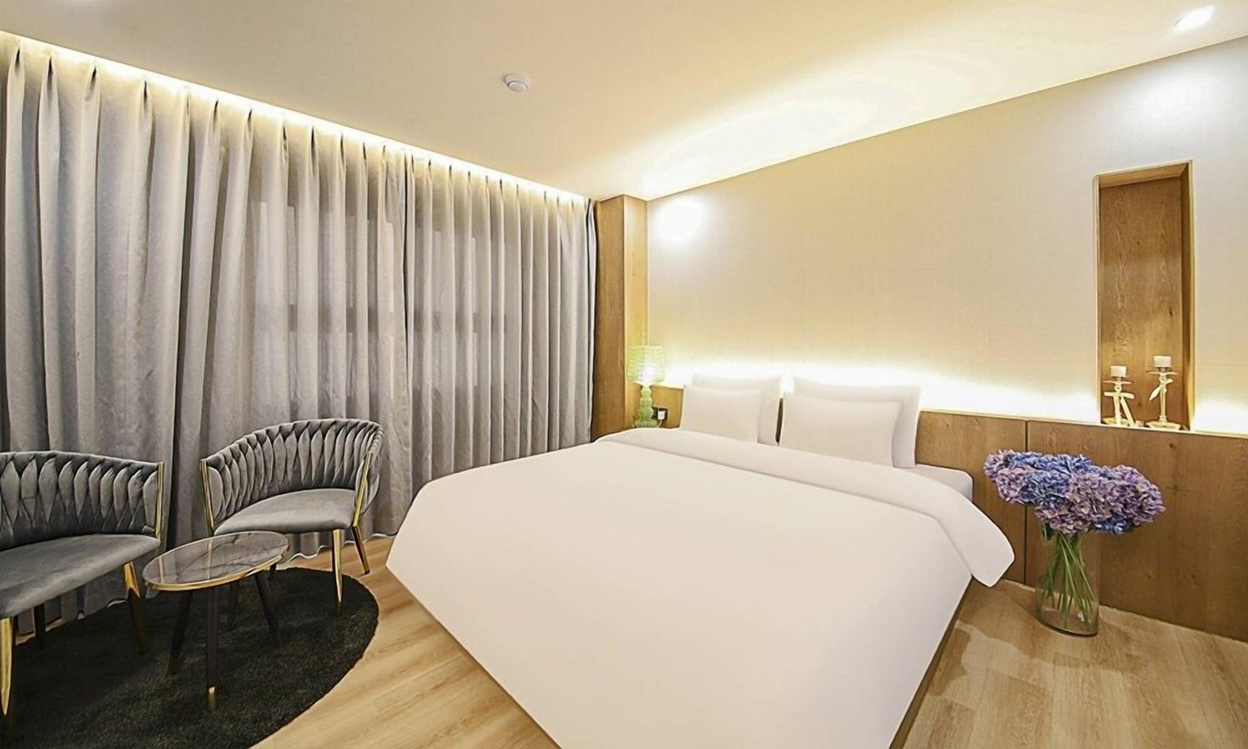 Quarto de hotel moderno com uma cama grande, duas poltronas estofadas, uma mesa redonda pequena, um espelho e um vaso com flores.