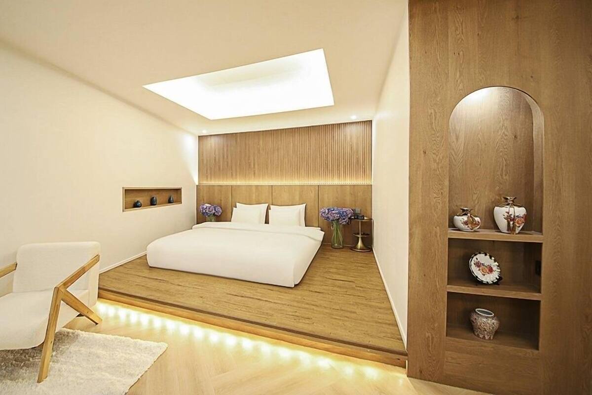 Quarto moderno com claraboia, cama, cabeceira de madeira, cadeira branca e aparador de madeira com objetos decorativos.