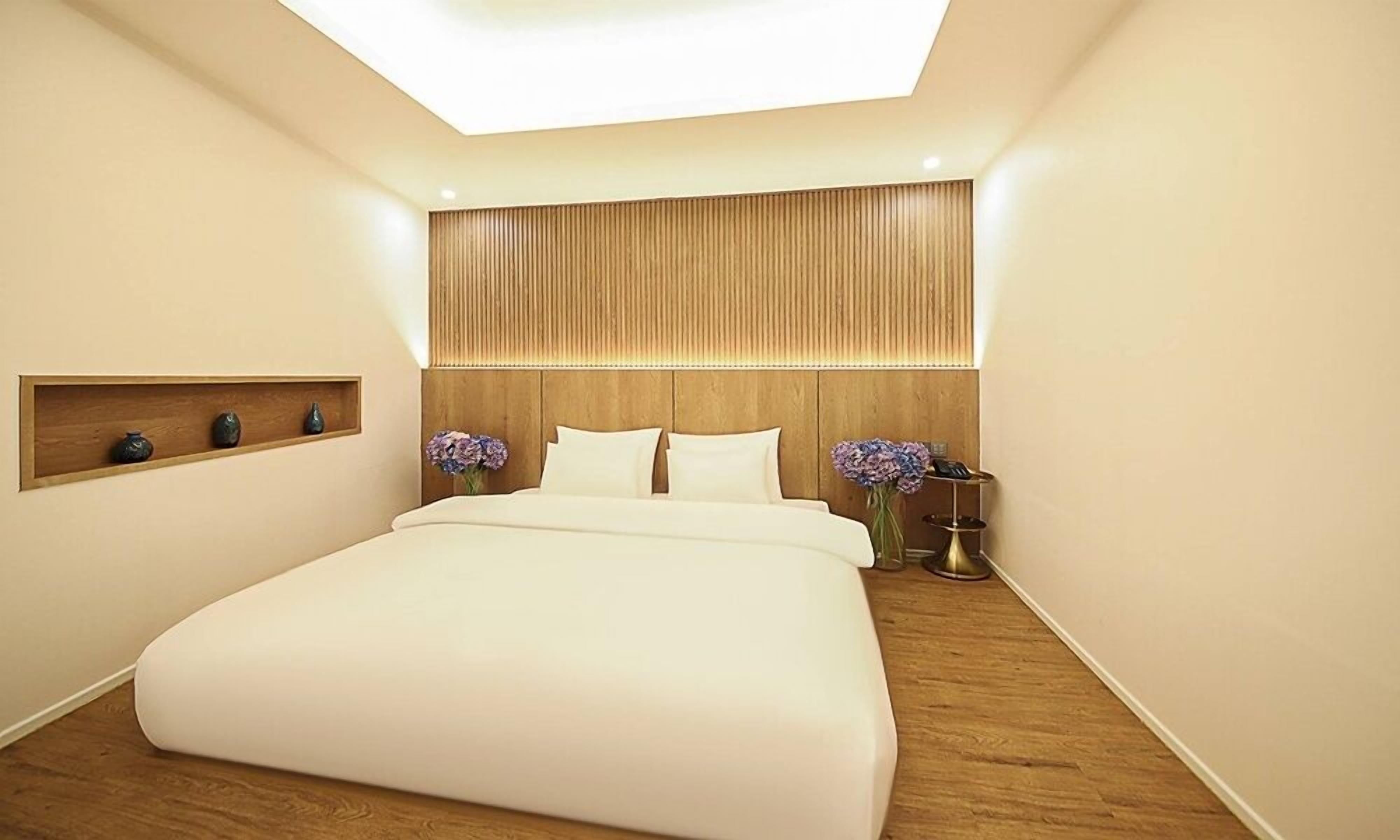A modern hotel room with a large bed, wooden headboard, built-in shelf, and a skylight.