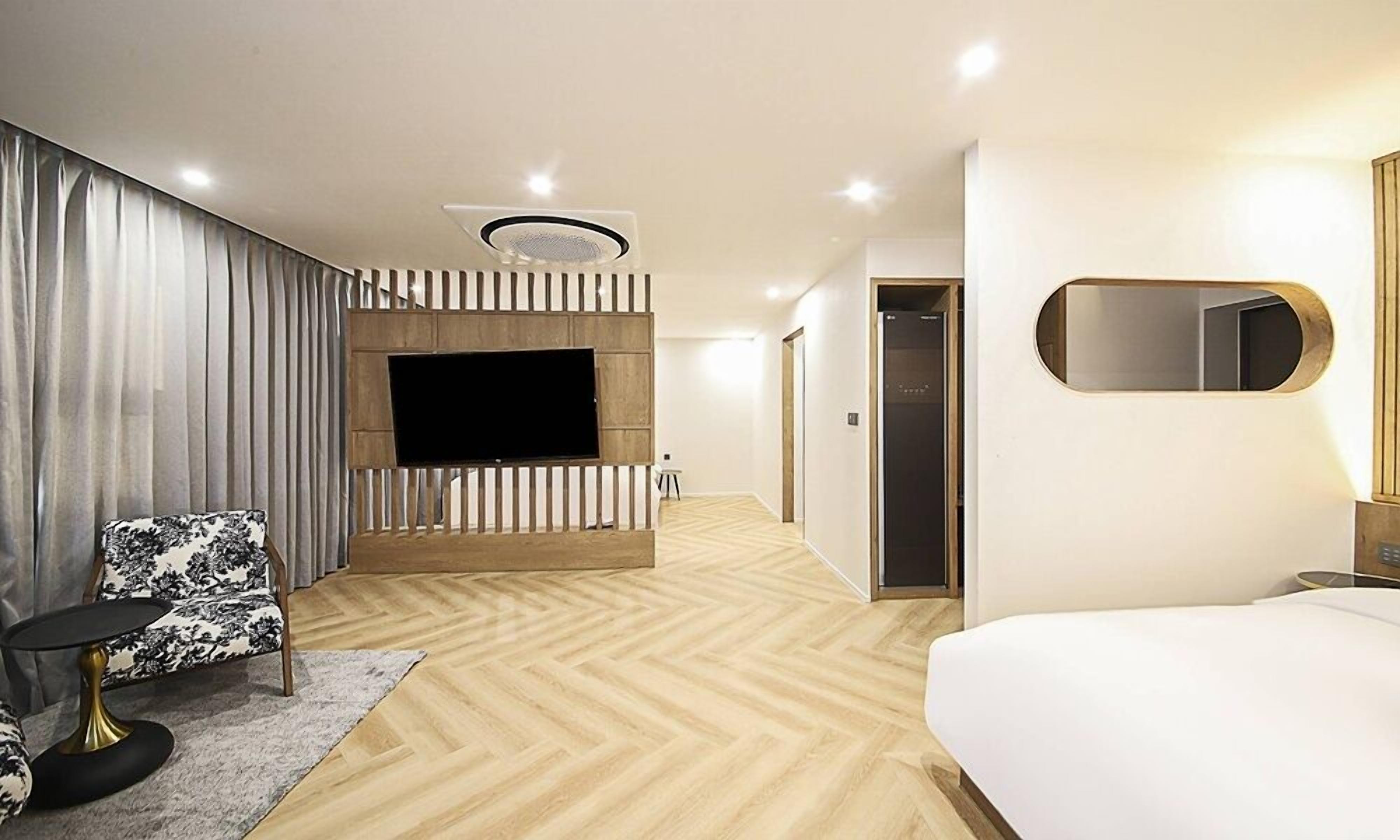 Quarto de hotel moderno com chão de madeira, uma cama, televisão fixada em uma parede revestida de madeira, uma mesinha de cabeceira e um espelho na parede.