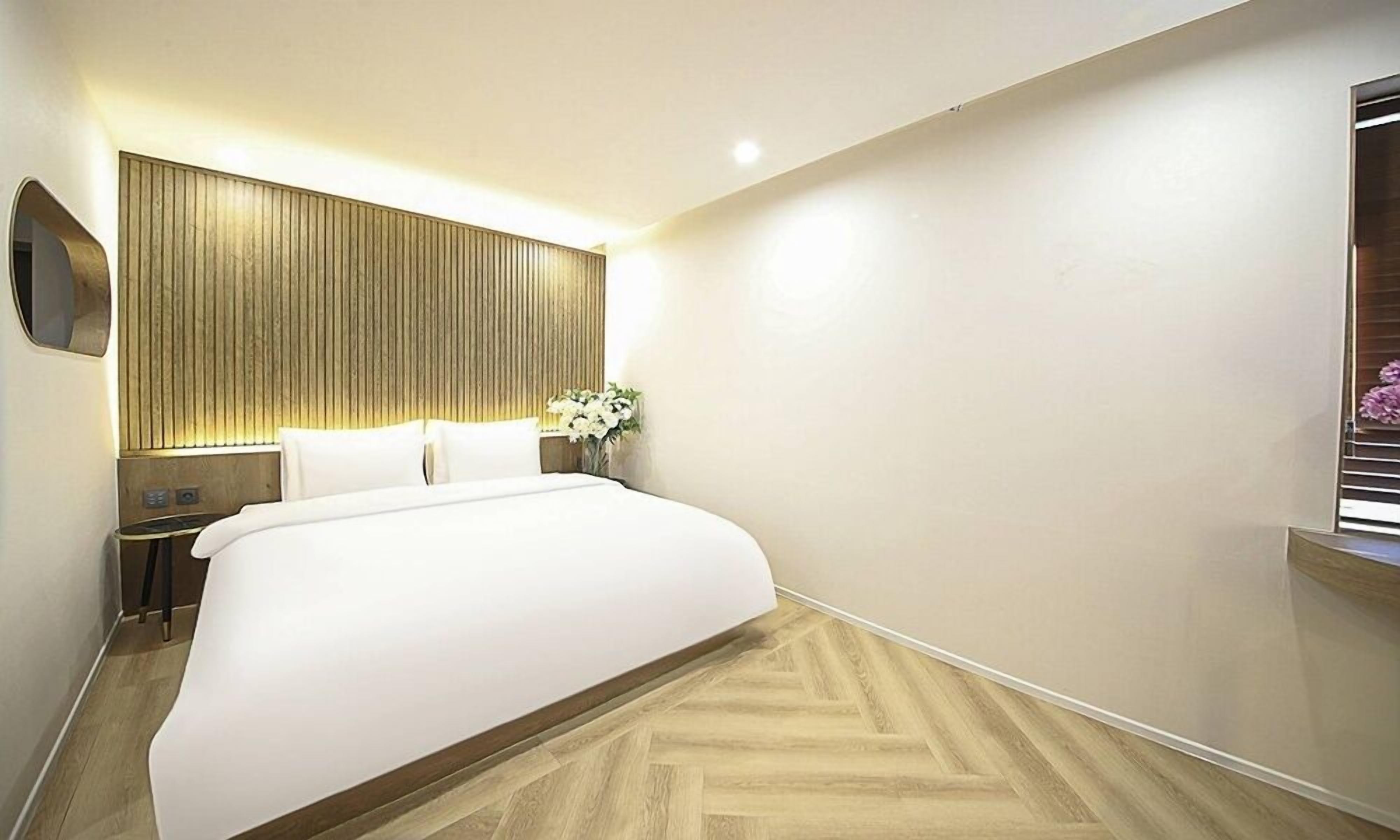 A modern hotel room with a large bed, a wooden headboard, a bedside table with a lamp, and a window with blinds.
