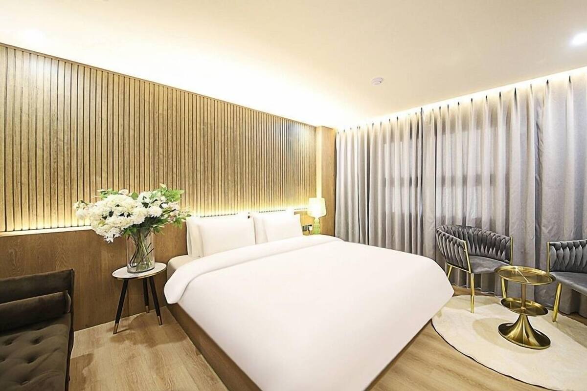 Quarto de hotel com uma cama grande, área de estar com duas poltronas e uma mesa redonda pequena, um vaso de flores e um painel de parede de madeira.
