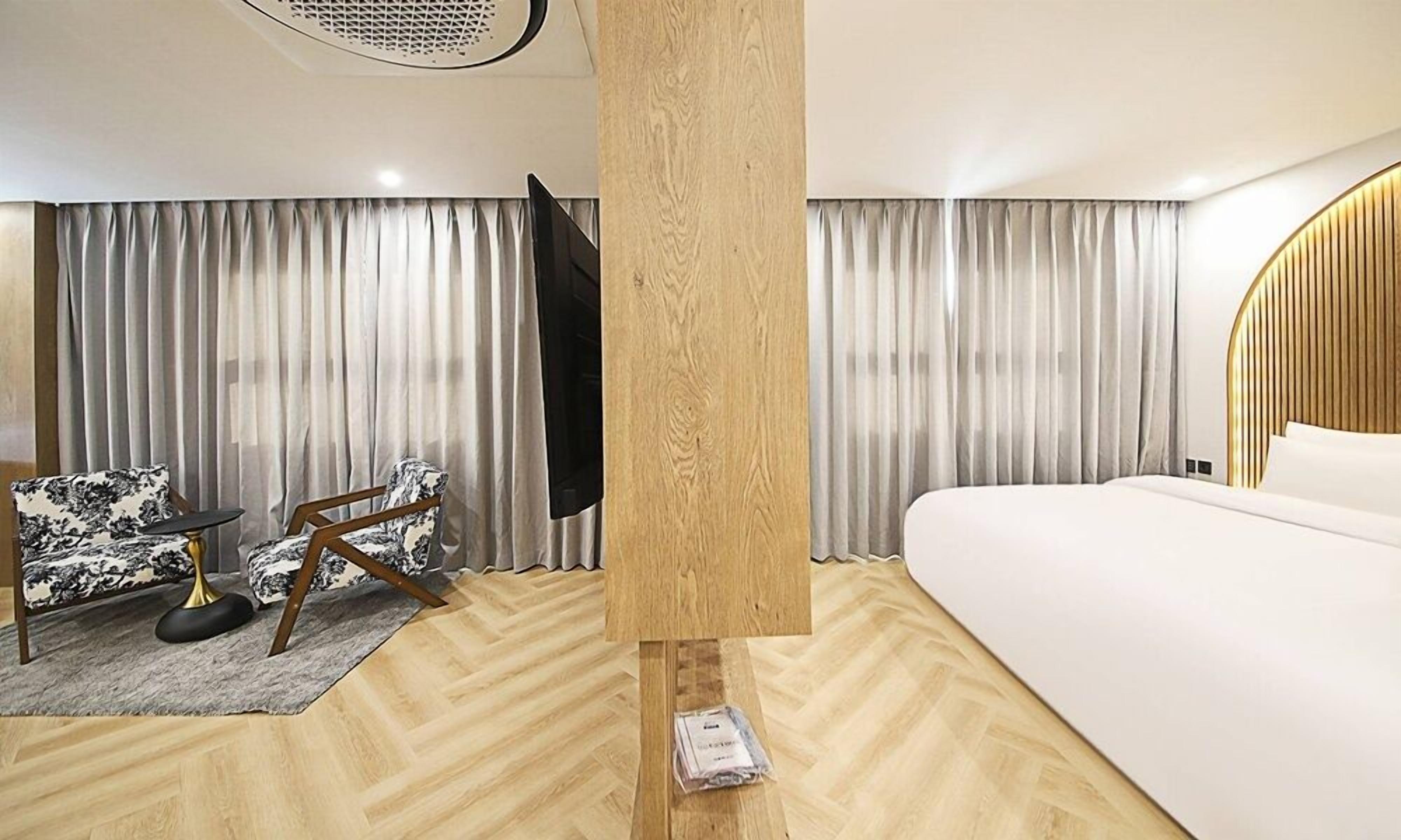 A modern hotel room with a wooden floor, a bed, a desk with a chair, and a large window with curtains.