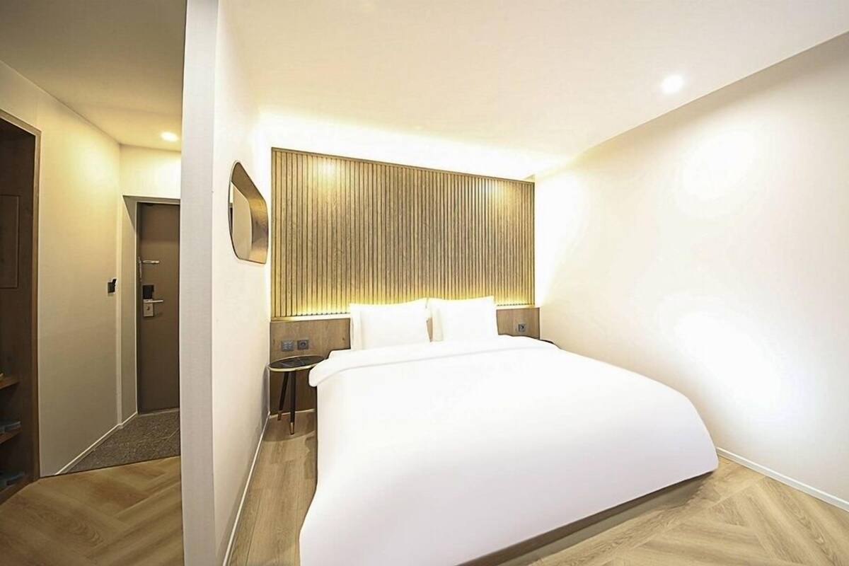 Quarto de hotel moderno com uma cama grande, uma mesinha de cabeceira, uma luminária na parede e uma porta.