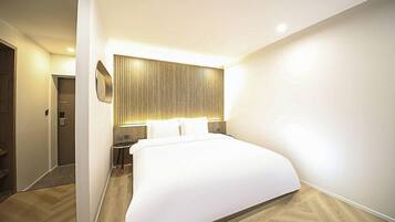 A modern hotel room with a large bed, a bedside table, a wall-mounted light, and a door.