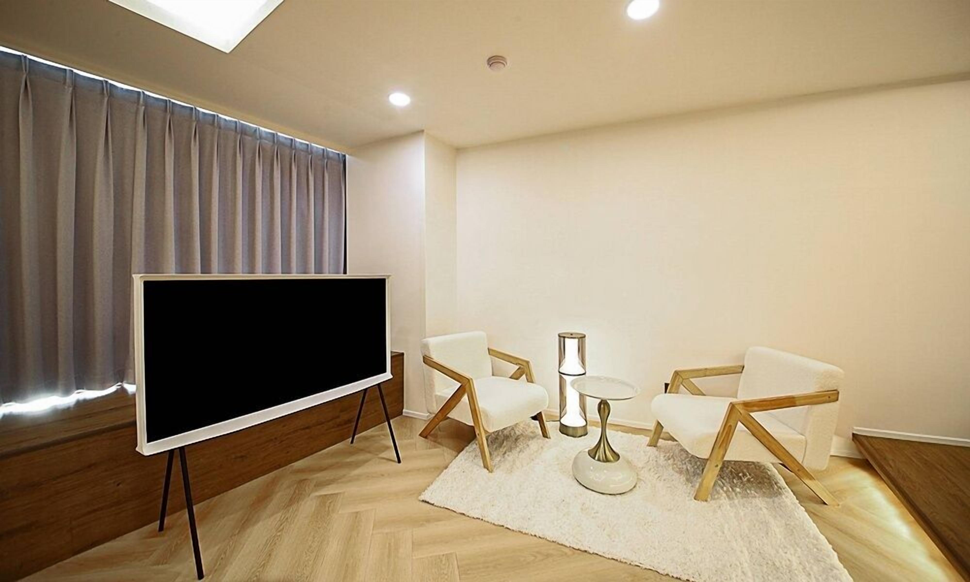 A modern room with a large flat-screen TV on a stand, two white chairs, a small round table, and a white rug.
