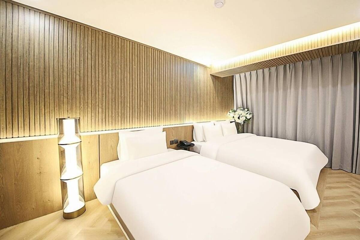 Quarto de hotel com duas camas, mesa de cabeceira com luminária, telefone e janela com cortinas.