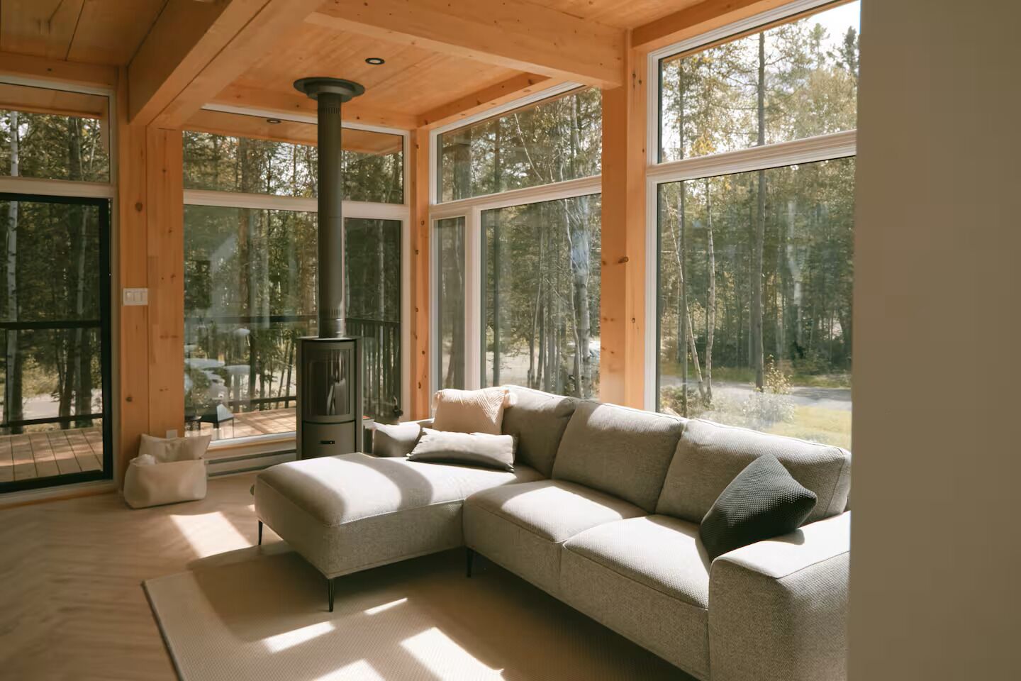 L’intérieur chaleureux d’un chalet avec un grand canapé d’angle, un poêle à bois et de grandes fenêtres donnant sur la forêt.