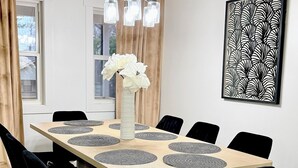 Una sala menjador amb taula rectangular, cadires negres, un vas amb flors blanques, una obra d'art mural amb disseny i un penjoll.