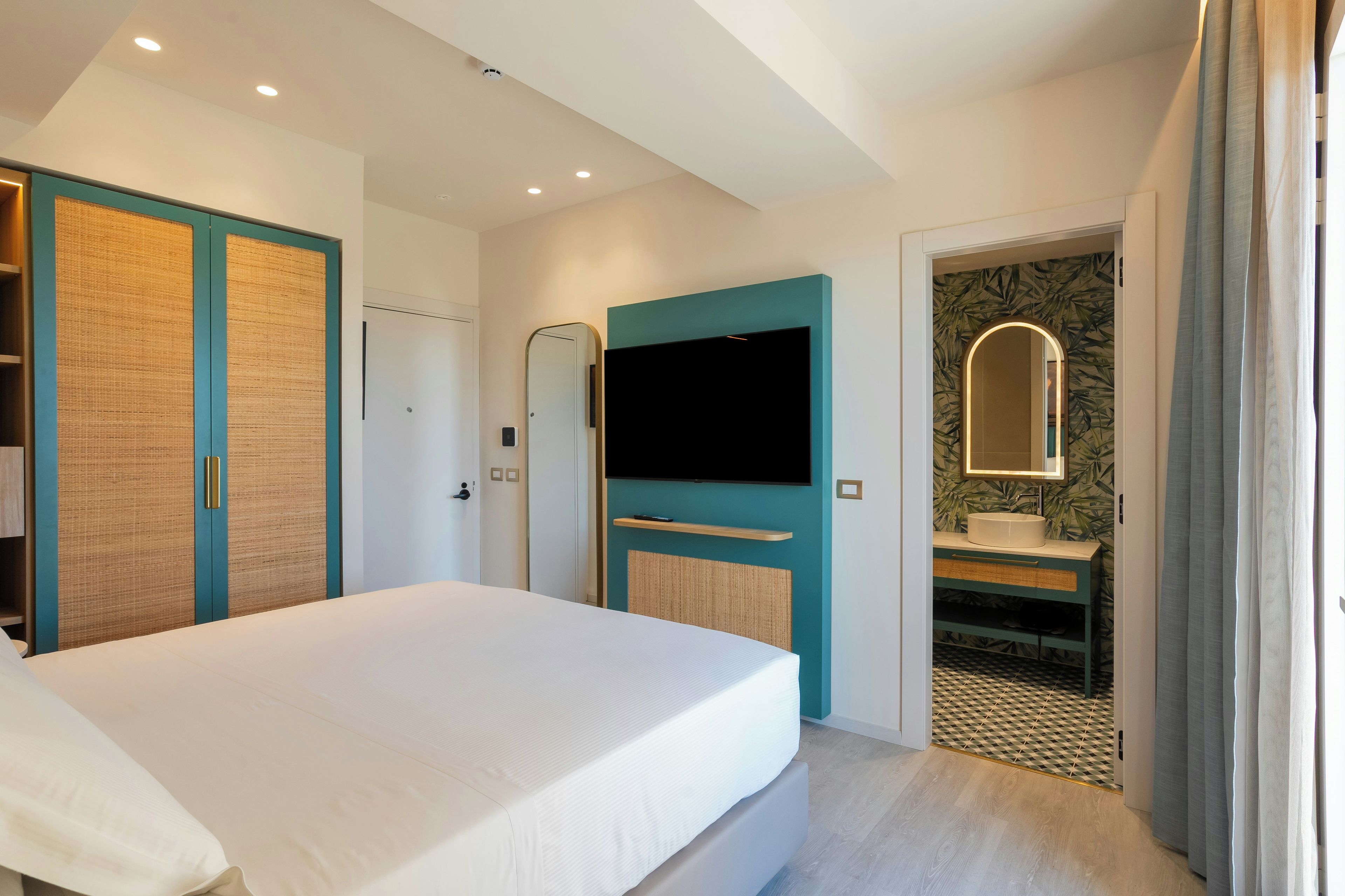 Habitación de hotel con cama, televisor montado en la pared, baño con espejo y puerta que da a otra habitación.