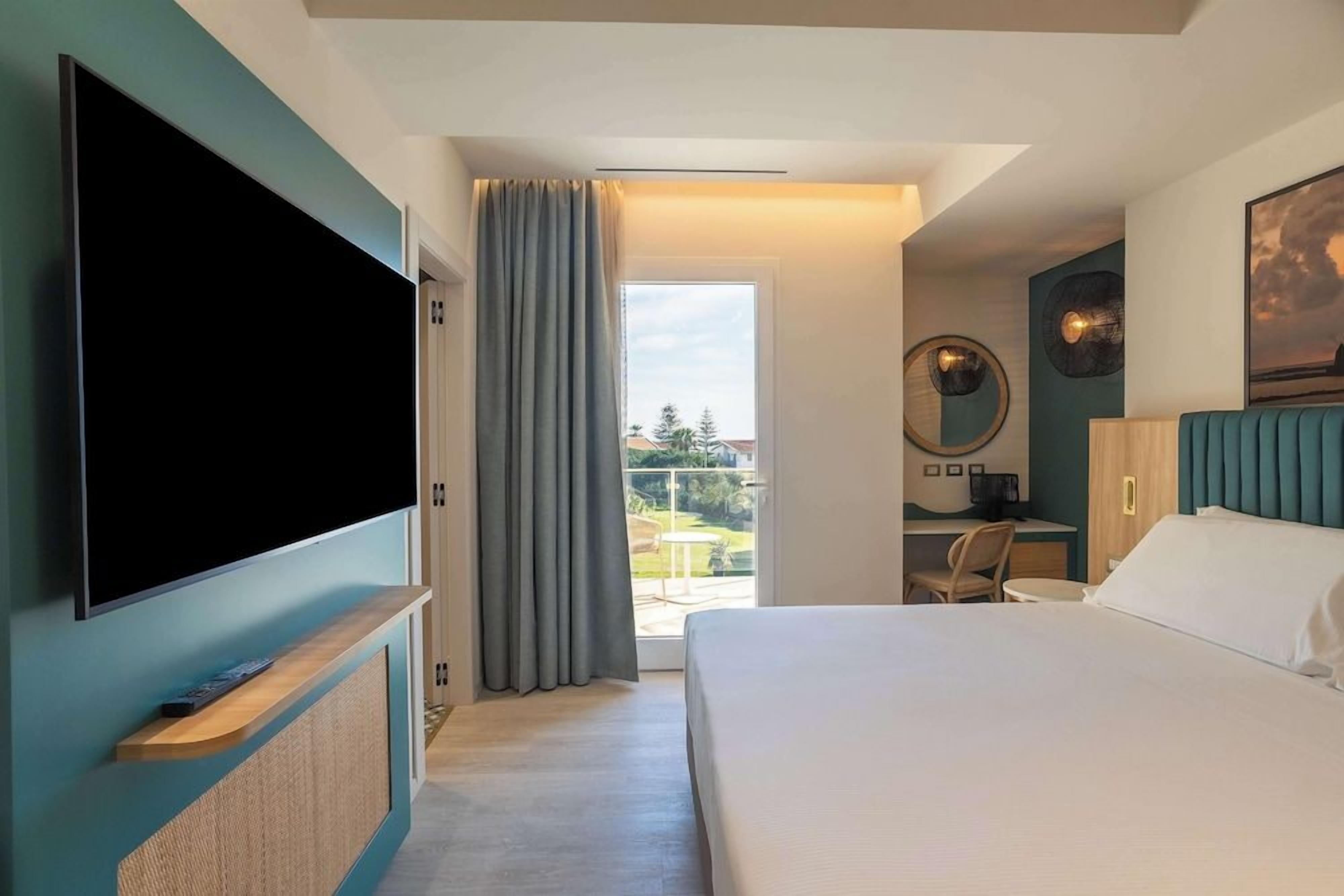 A hotel room with a bed, a TV, a desk with a chair, and a view through a door to an outdoor area.
