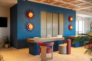 Una reception con una TV a schermo piatto montata a parete, una targa con il nome dell'hotel e un'area salotto con una poltrona e un poggiapiedi rossi.