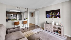 A modern living room with a sofa, a coffee table, and a TV mounted on the wall. There is a kitchen area with a microwave, refrigerator, and a small bar counter with two chairs. - FTL 1BR Near Beach | Pool + Free Parking (Fort Lauderdale)