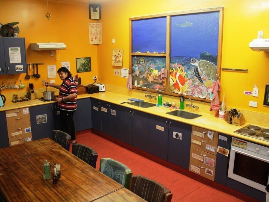 Une cuisine où une personne est en train de cuisiner, une fenêtre avec un tableau, et un coin repas avec une table et des chaises.