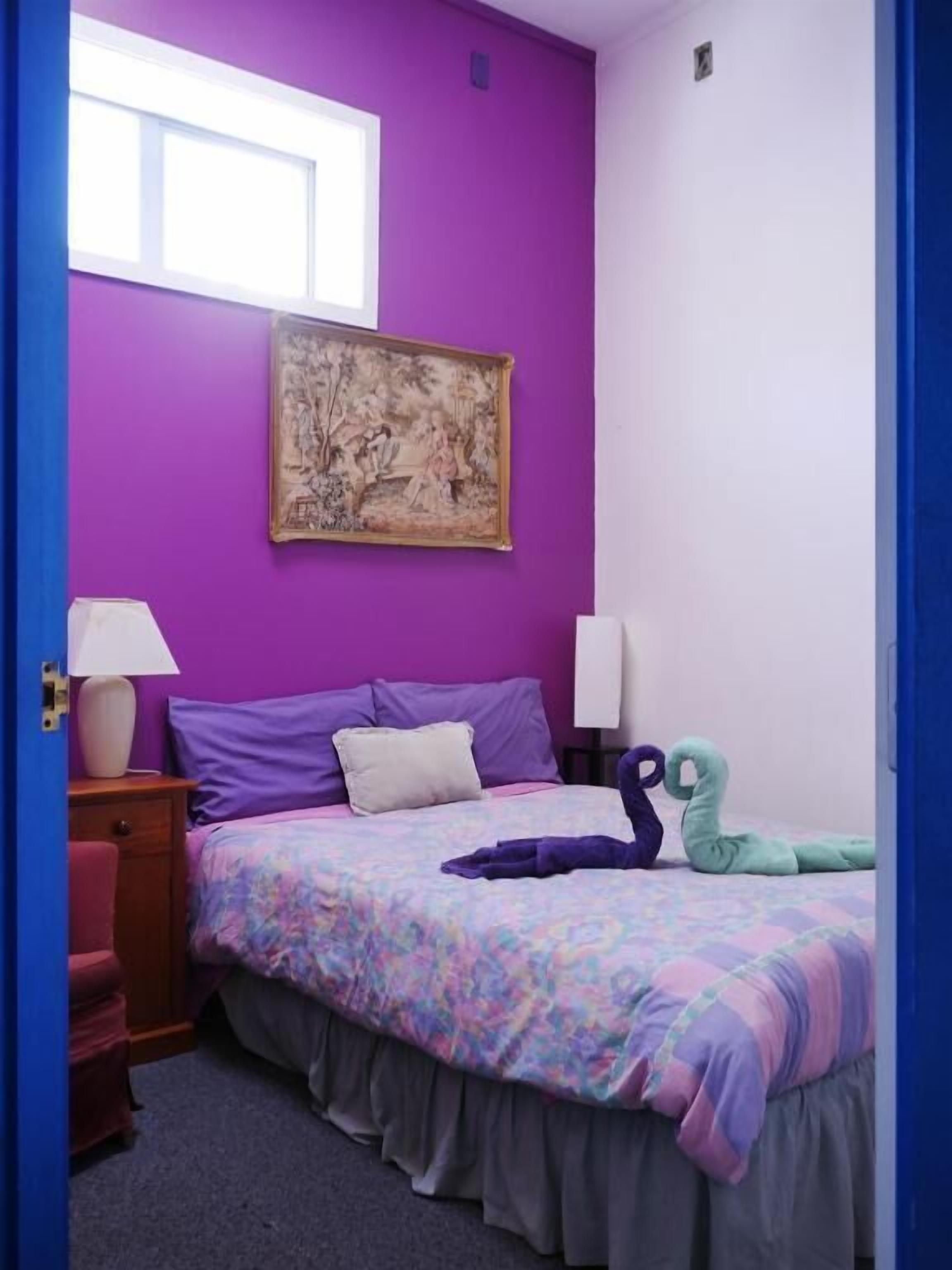 Une chambre avec un mur violet, un lit recouvert d’une couvre-lit à motifs floraux, deux lampes de chevet, une table de chevet et une fenêtre.