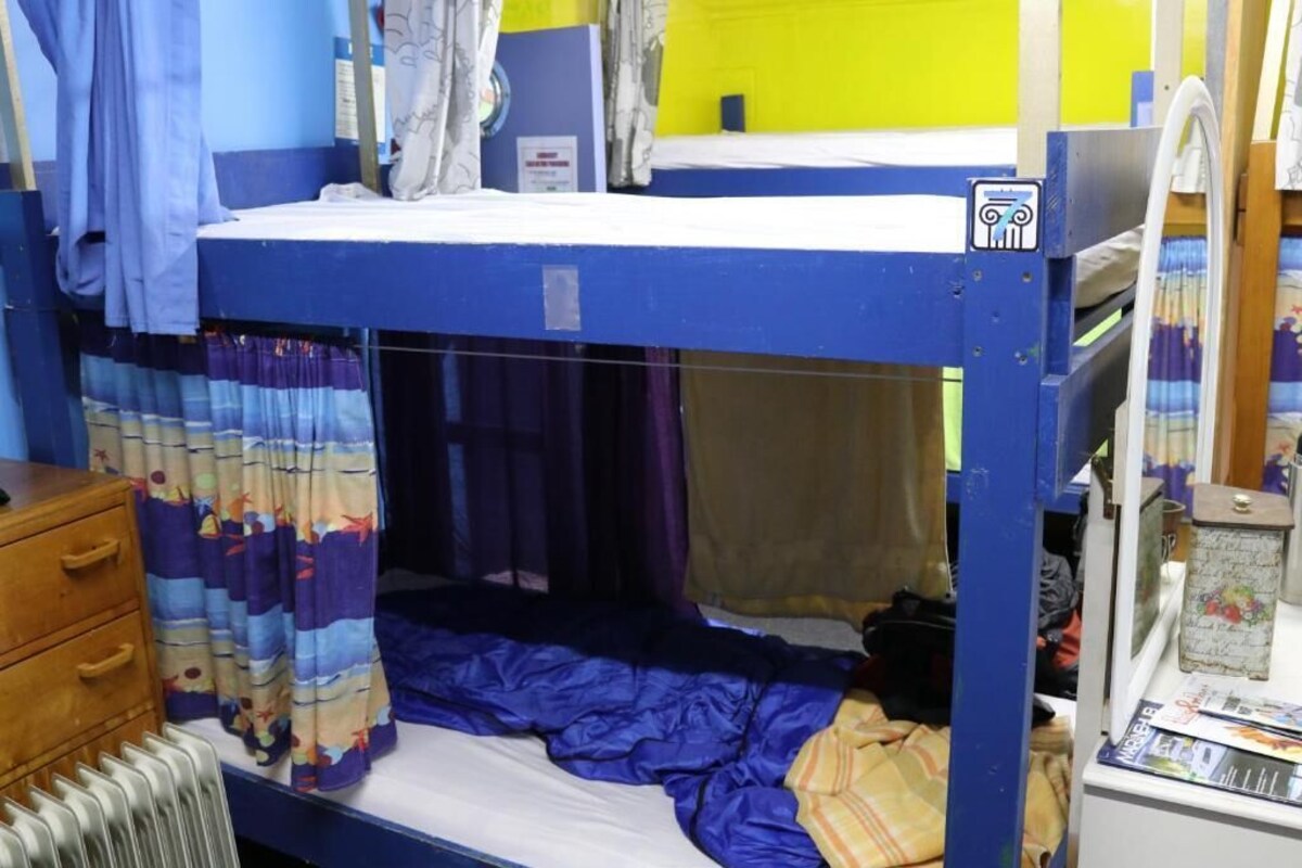Una cama litera con una litera superior azul, una litera inferior con cortina, un aparador de madera y un radiador.