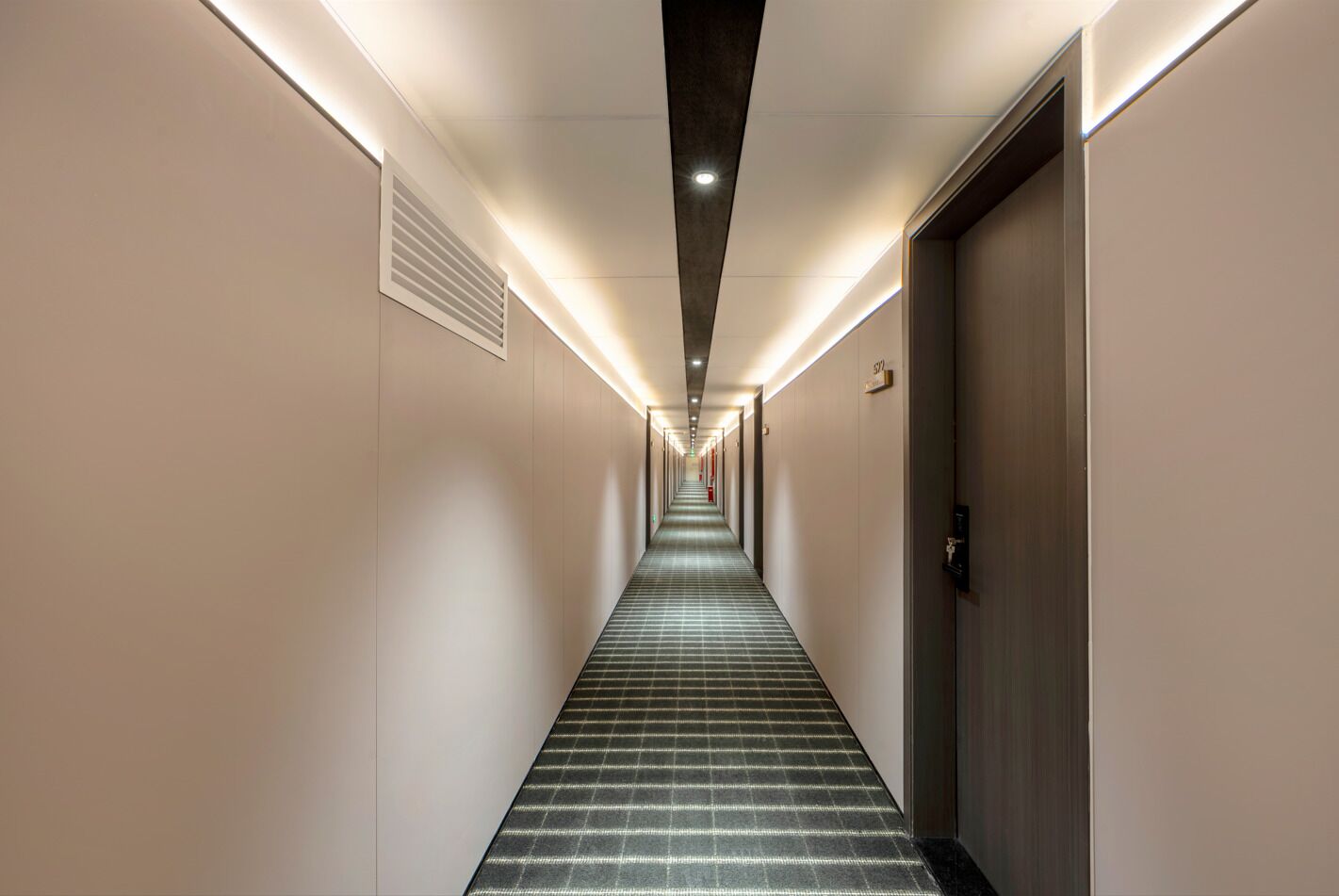 A long, narrow hotel corridor with a checkered carpet, recessed lighting, and doors on both sides.