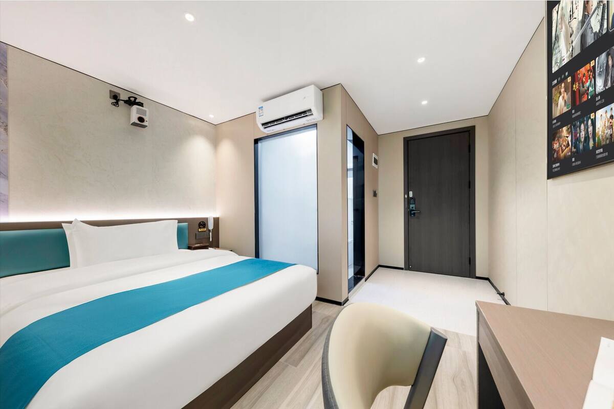 Ein modernes Hotelzimmer mit einem großen Bett, einem Schreibtisch und einem Stuhl. An der Wand ist eine Klimaanlage angebracht und rechts befindet sich eine Tür.