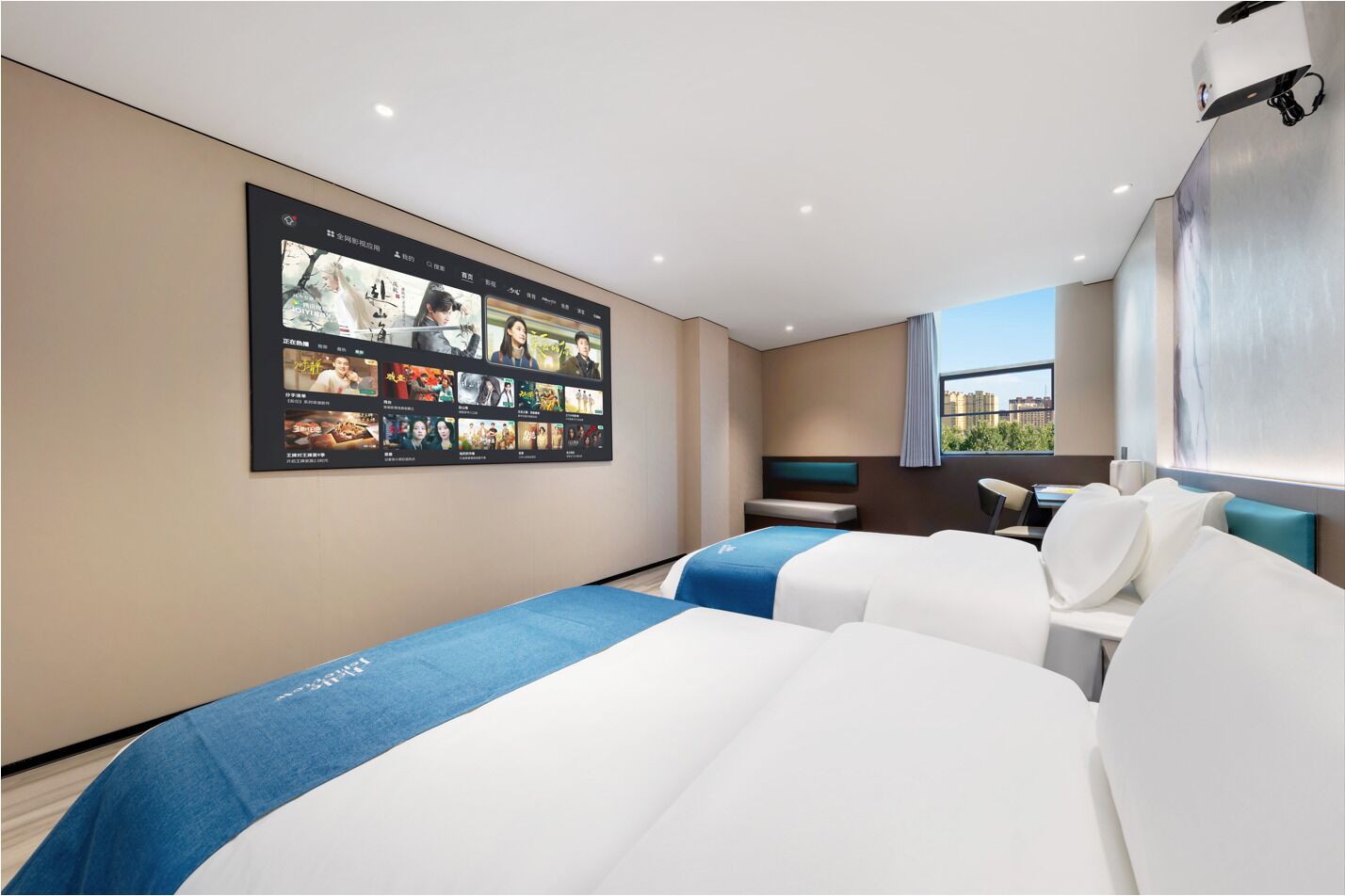 Habitación de hotel con una gran pantalla digital mostrando carteles de películas, una cama con colcha azul y blanca, un escritorio con una silla y una ventana con vista a la ciudad.