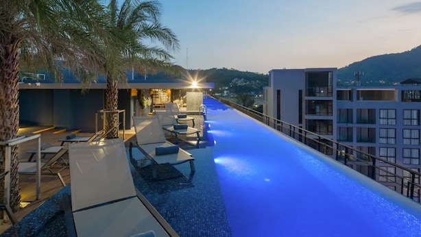 A rooftop infinity pool with lounge chairs and palm trees, overlooking a cityscape at dusk. - Stylish 2BR Condo in Citygate Phuket – Walk to Beach, Clean & Breezy Stay (Kamala)