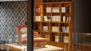 A cozy reading nook with a leather armchair and ottoman, a wooden bookshelf filled with books, and a patterned wallpaper backdrop. - Stylish 2BR Condo in Citygate Phuket – Walk to Beach, Clean & Breezy Stay (Kamala)
