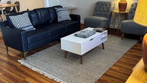 Uma sala de estar moderna com um sofá azul escuro, uma mesa de centro branca e uma poltrona cinza. Há obras de arte emolduradas na parede e uma luminária em uma mesinha lateral.