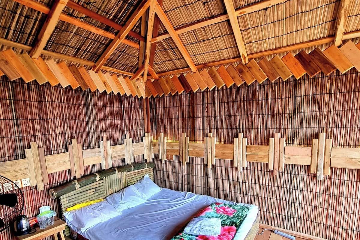 Una habitación con una cama, piso de madera, paredes de bambú y techo de paja.