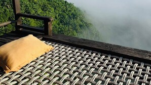 Eine Holzterrasse mit gewebtem Boden, mit Blick auf einen üppigen grünen Wald und ein von Nebel bedecktes Gebirge.