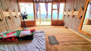 Ein Zimmer in einer Holzhütte mit einem Bett, einem Standventilator und Blick ins Freie.