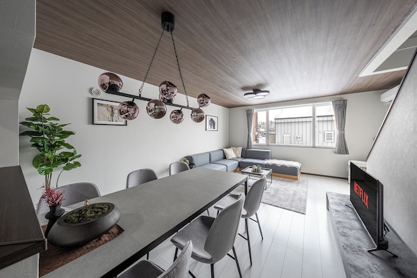 A modern living room with a dining area, a grey sofa, a flat-screen TV, and a large window. - HILLS NISEKO A (Niseko)