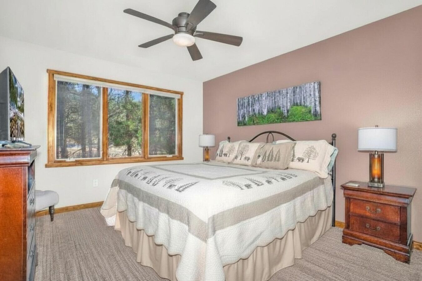 Ein Schlafzimmer mit Bett, Deckenventilator, Fenster mit Blick auf Bäume, einem Bild an der Wand und einem Nachttisch mit Lampe.
