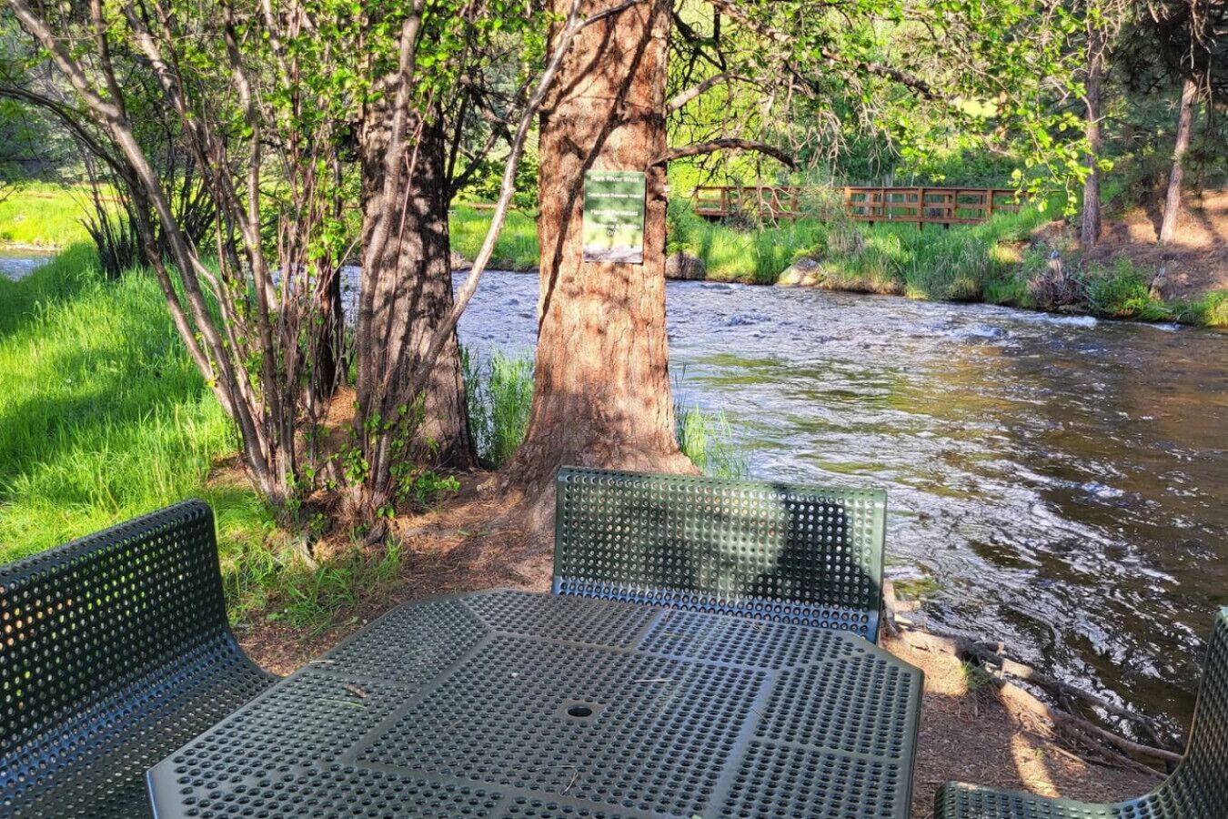 Une table de pique-nique et des chaises au bord d’une rivière, avec un pont en bois en arrière-plan.