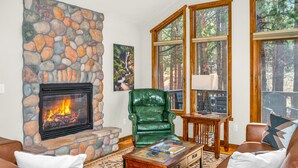 A living room with a stone fireplace, leather armchairs, a coffee table, and large windows with a forest view.