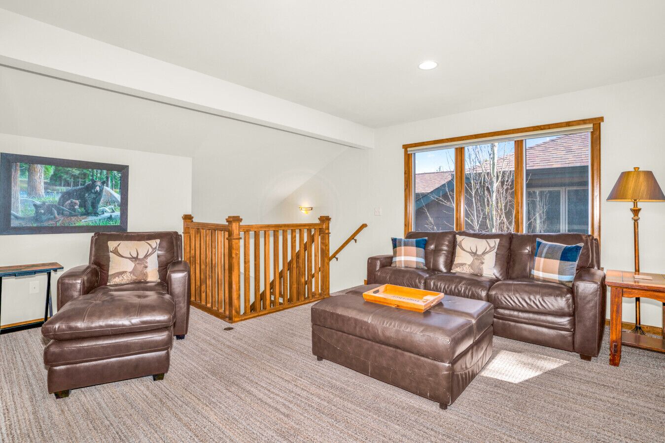 A living room with a brown leather sofa, a brown leather armchair, a wooden staircase, and a large window with a view of trees.