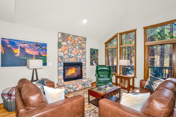 A living room with a stone fireplace, leather sofas, a coffee table, and large windows with a forest view.
