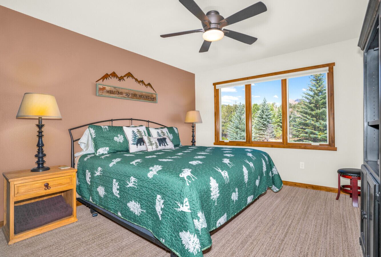 A bedroom with a bed, a nightstand, a lamp, a window with a view of trees, and a ceiling fan.