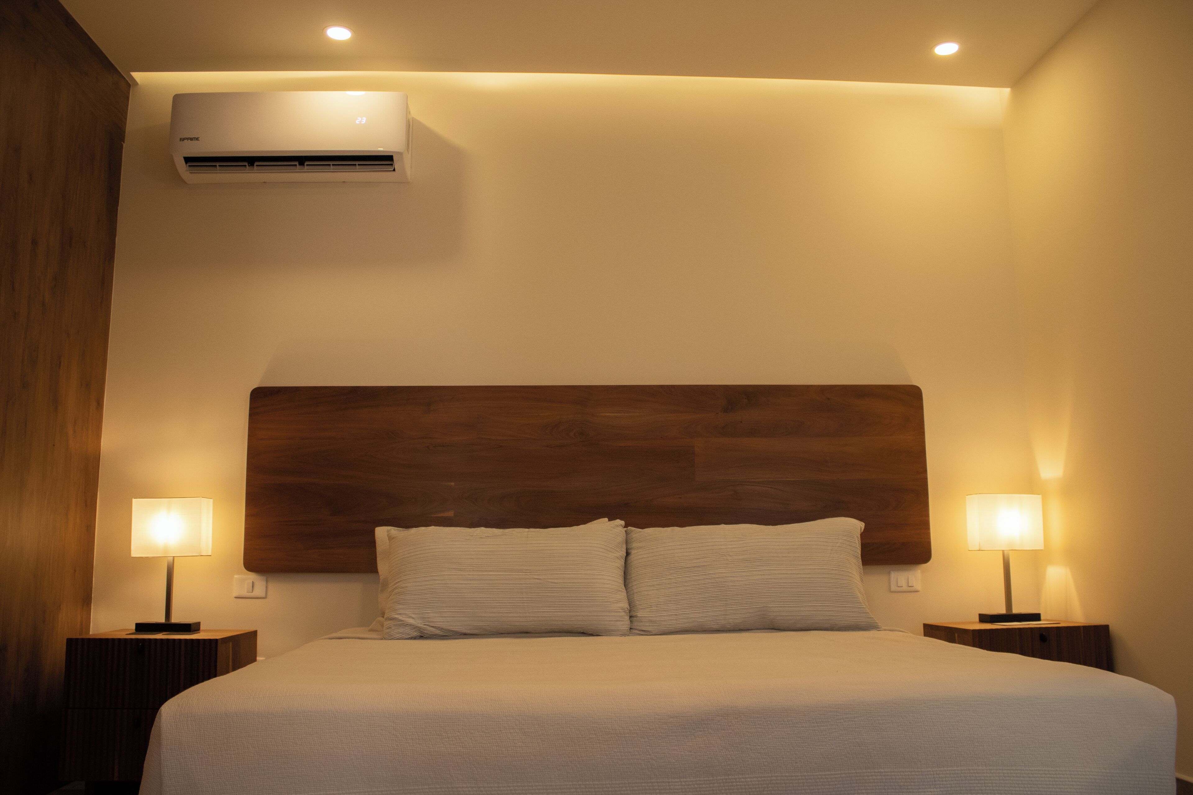 A neatly made bed with a wooden headboard, two bedside lamps, and an air conditioning unit on the wall.