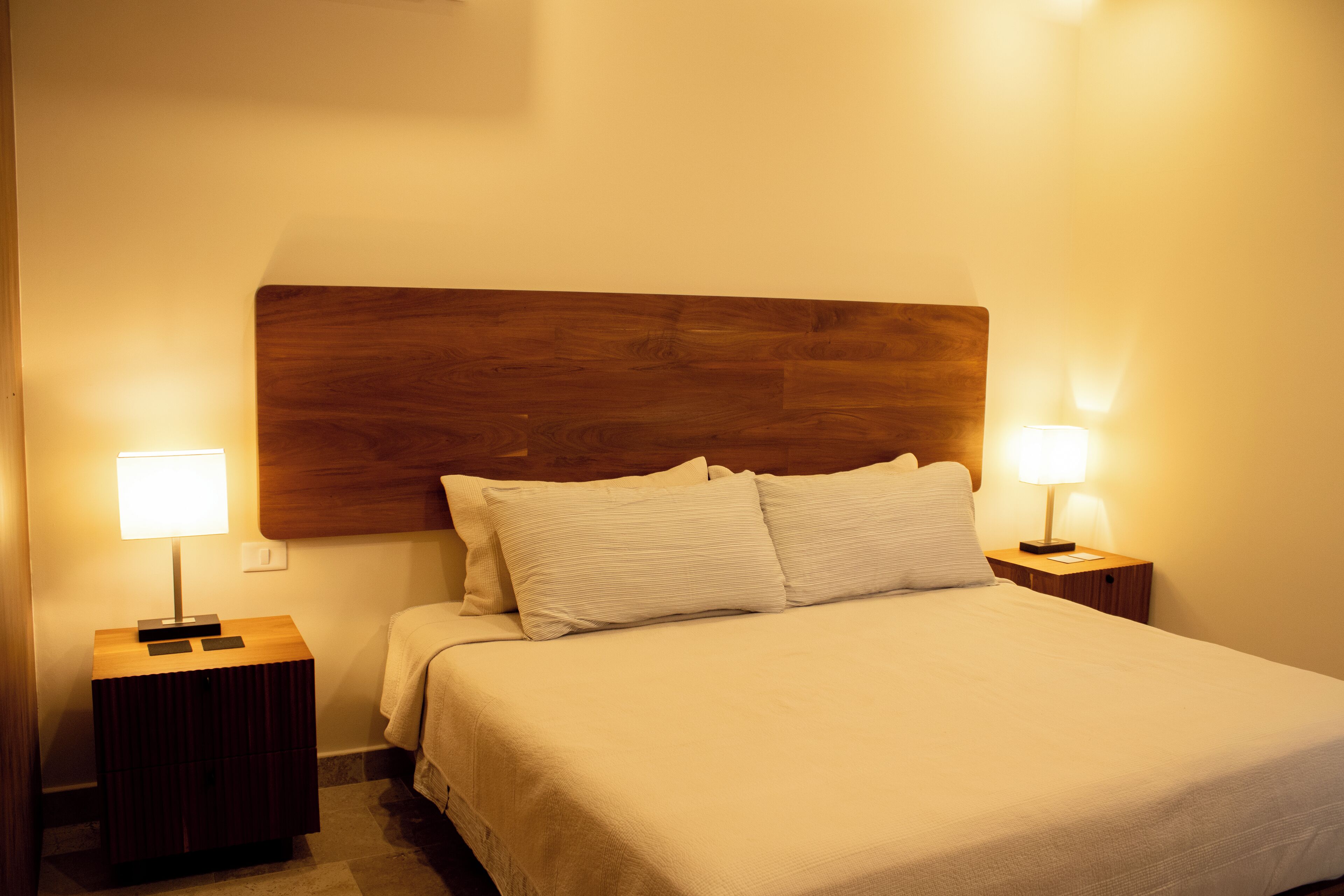 Een net opgemaakt bed met een houten hoofdbord, twee bedlampjes en een klein nachtkastje.