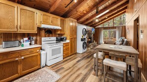 A log cabin interior with wooden cabinets, a kitchen area, a dining table with benches, and a large window providing a view of the outdoors. - Mama Bear's Cozy Cabin - Fireplace, Central Heat, Spacious Deck (Sugarloaf)
