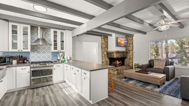 Une cuisine moderne avec des meubles blancs, une cheminée en pierre et un coin repas agrémenté d’un canapé gris et d’un tapis bleu.