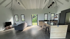 A spacious living area with a grey sofa, a TV, and a kitchen with a wooden island and stools. - Villa le Paradis Secret: Sea view, river access. (Goyave)