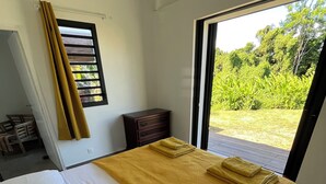 A bedroom with a bed, a wooden dresser, and a view of greenery outside. - Villa le Paradis Secret: Sea view, river access. (Goyave)
